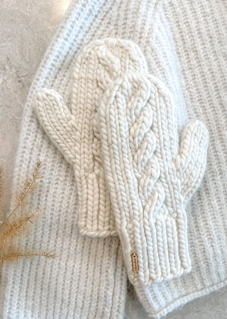Pair of cream-colored knitted mittens on a textured surface with gold leaves.