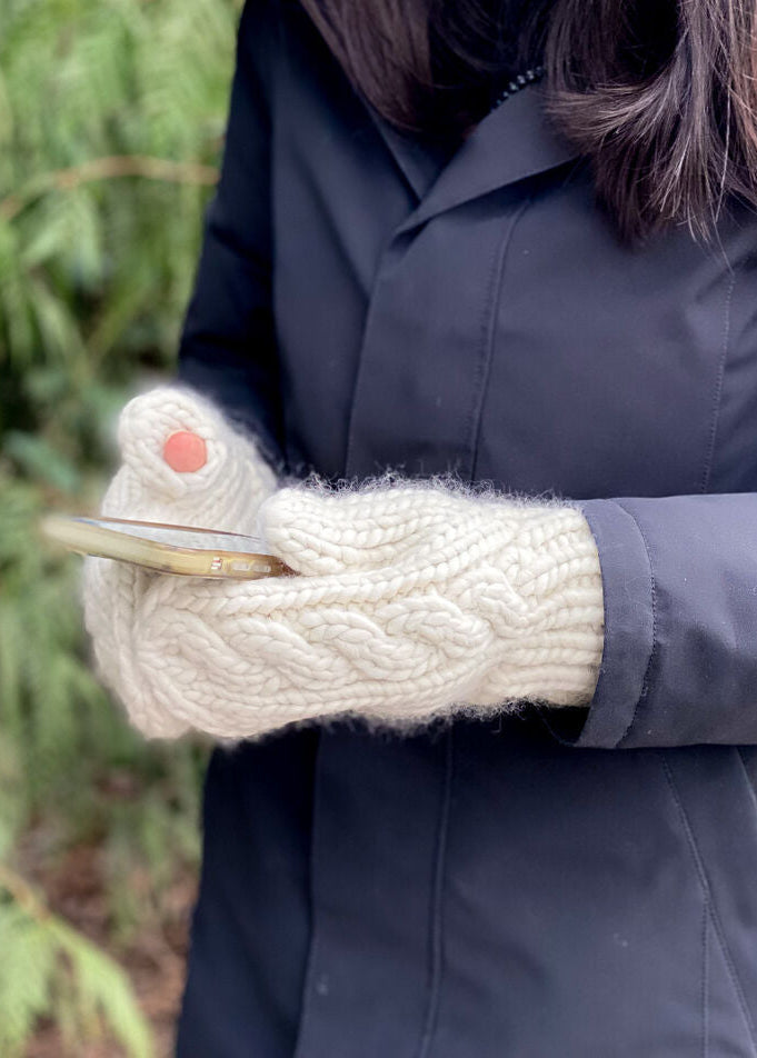Person wearing a dark coat and white knitted mittens holding a phone with a blurred natural background