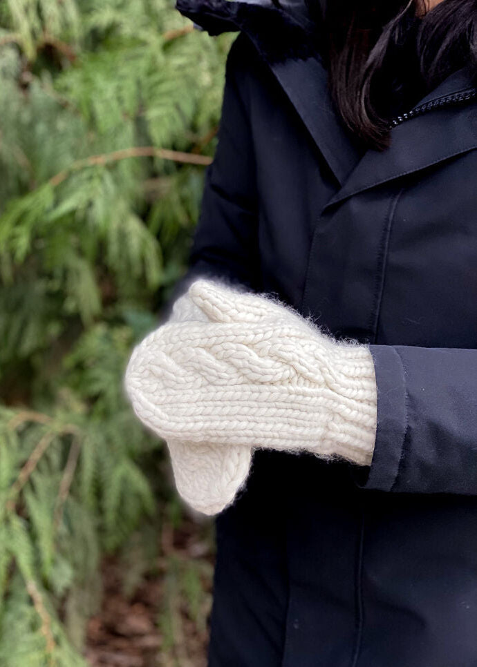 Person wearing a dark coat and white knitted mittens with a blurred green forest background