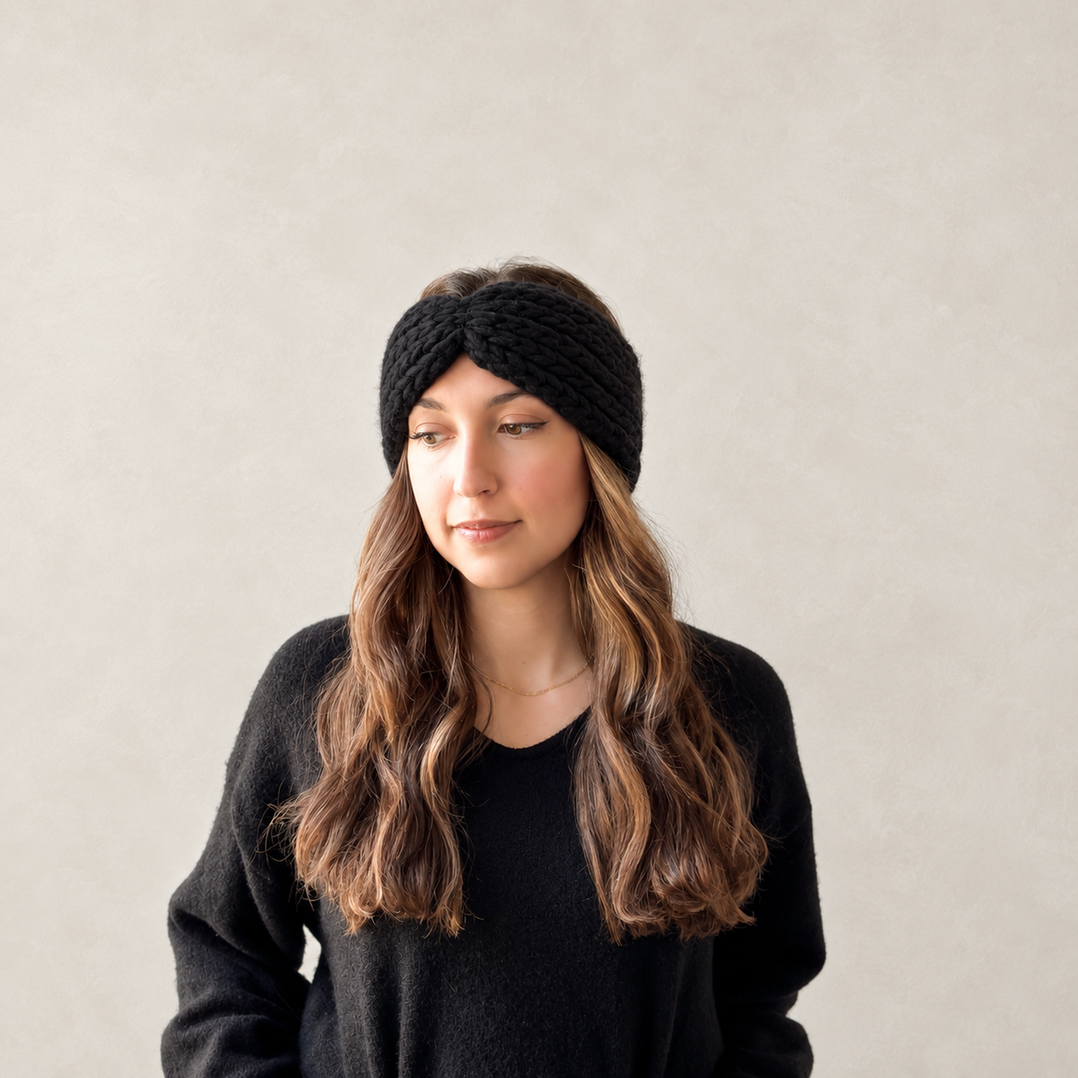 A woman with long wavy brown hair wearing a black chunky knit headband and a black knit sweater, photographed against a plain light beige wall.