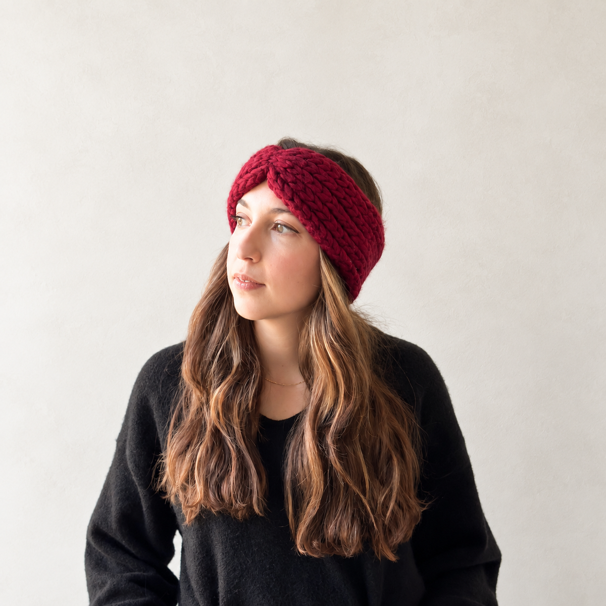 A woman with long wavy brown hair wearing a chunky, textured burgundy knit headband and a black sweater, looking to the side against a plain light gray wall.