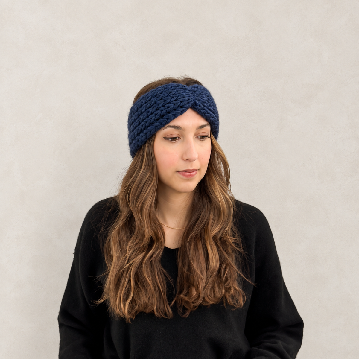 A woman with long wavy brown hair wearing a thick navy blue knitted twisted headband and a black sweatshirt, standing against a textured light gray wall.