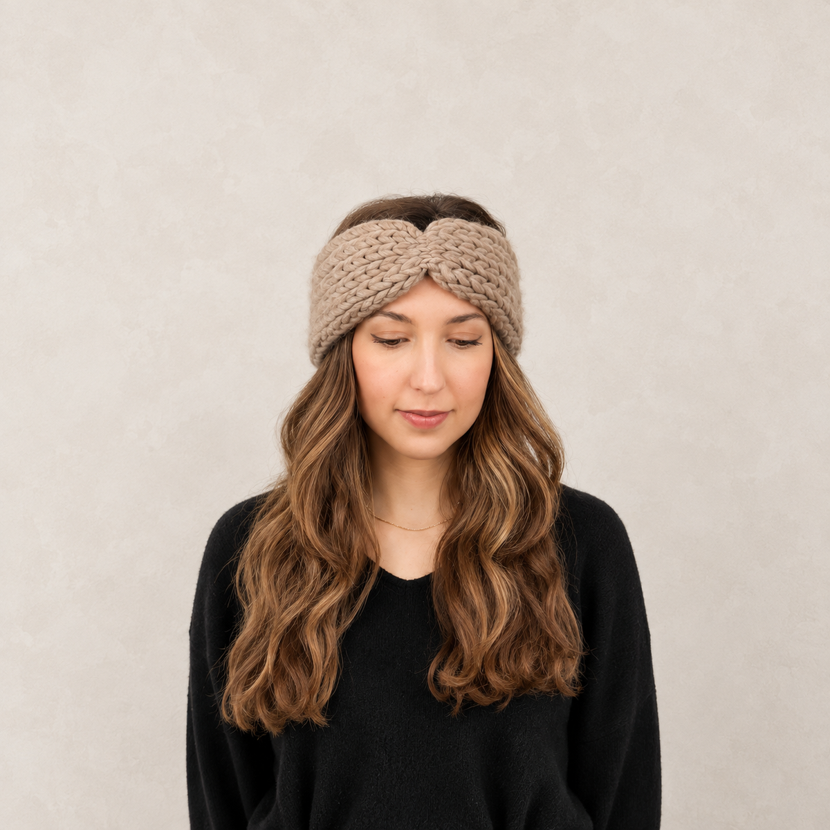 A woman with long wavy brown hair wearing a chunky knit twisted headband in taupe and a black sweater, photographed against a neutral textured beige background.