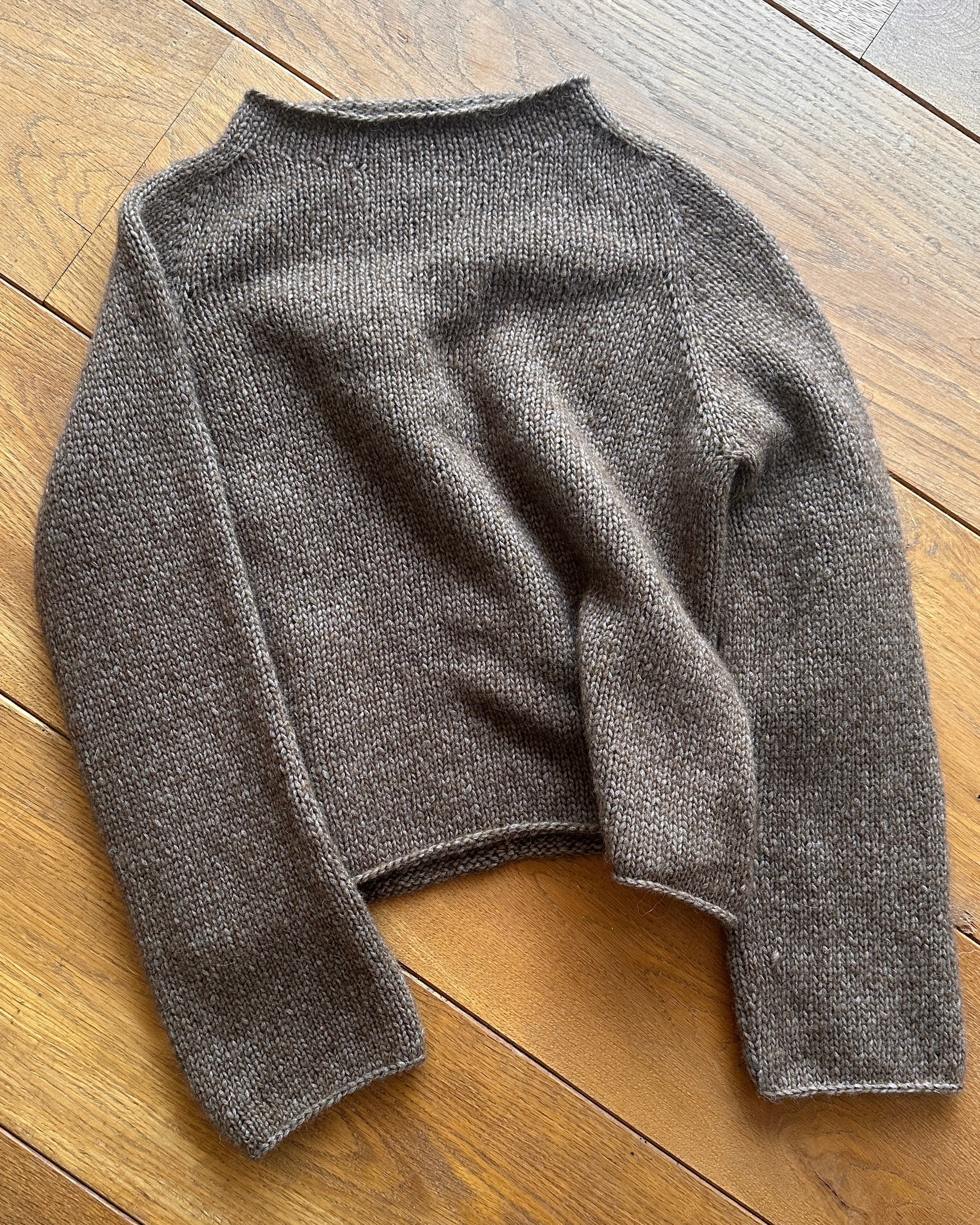 A brown knit sweater with a crew neck and long sleeves, laid flat on a wooden floor.