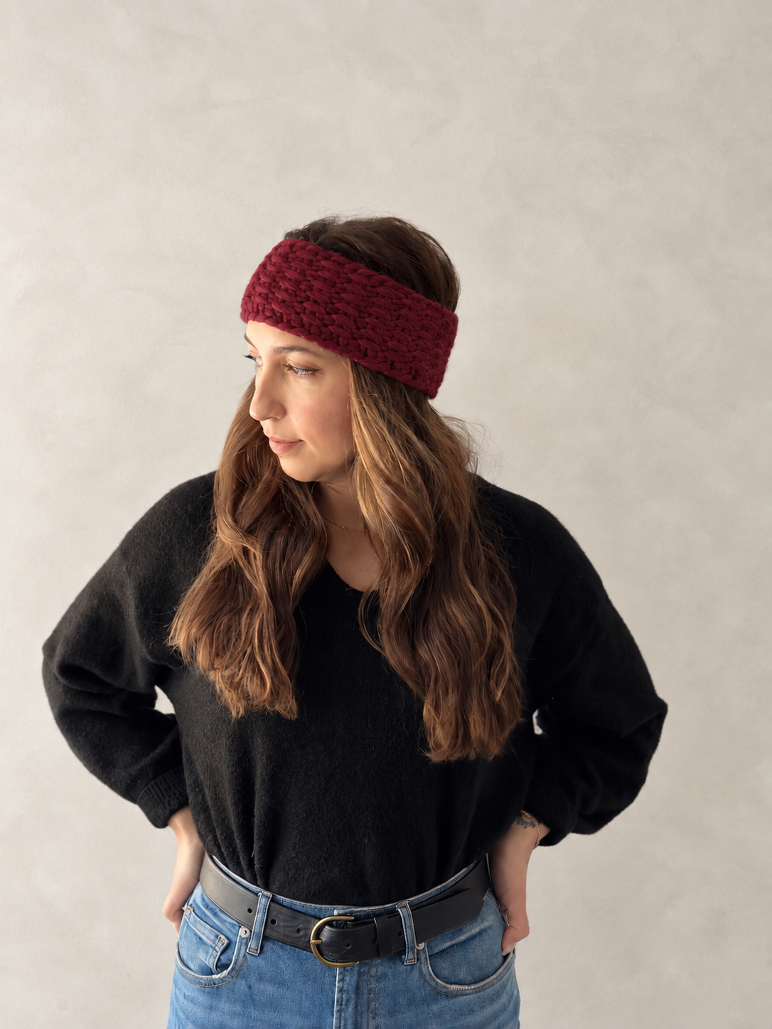 A woman wearing a chunky, dark red crocheted headband, a black long-sleeve top, and blue jeans, standing against a neutral textured beige wall.