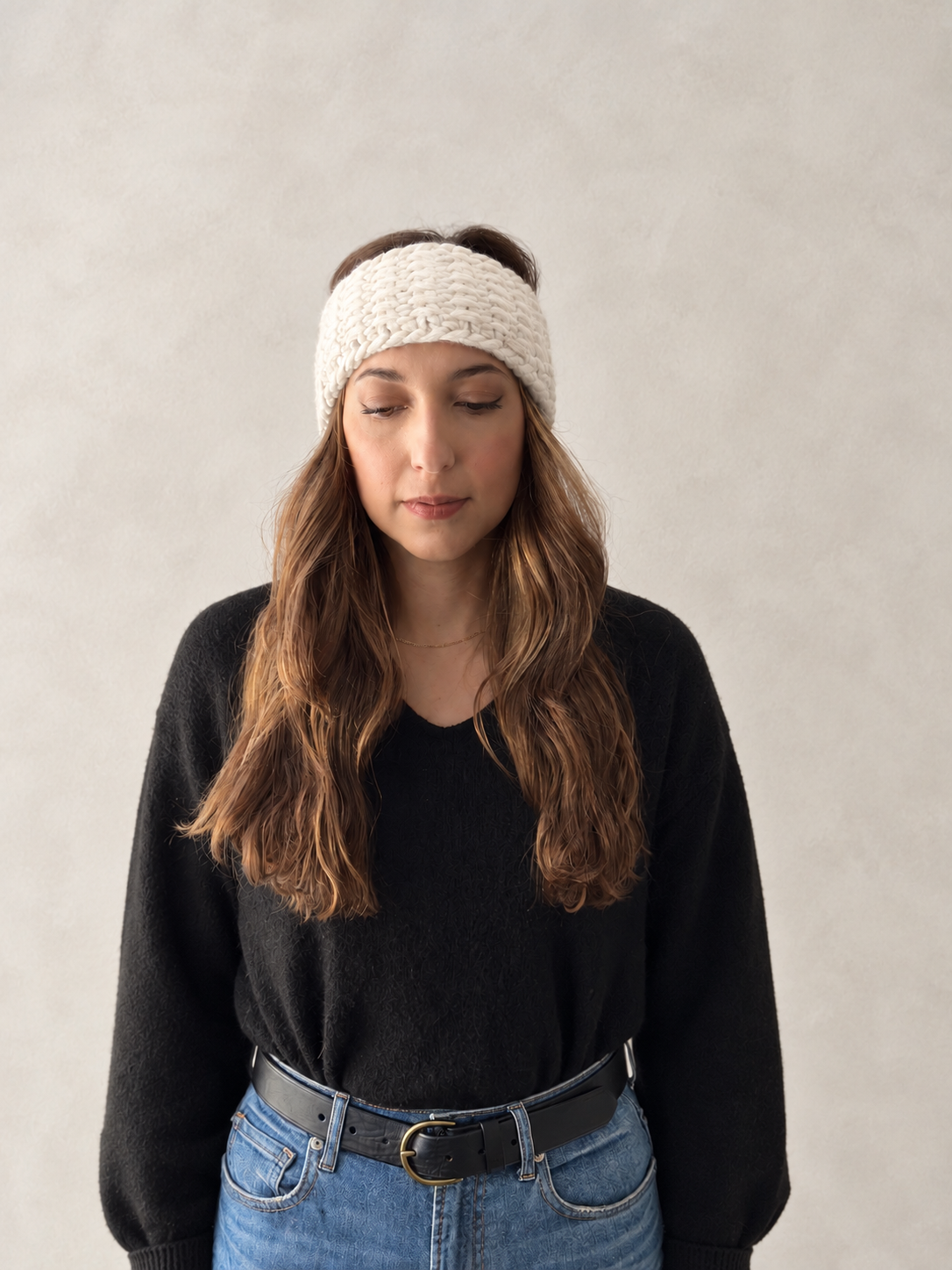A woman wearing a cream-colored, chunky-knit headband, a black knit sweater, and blue jeans, standing against a neutral textured gray wall.