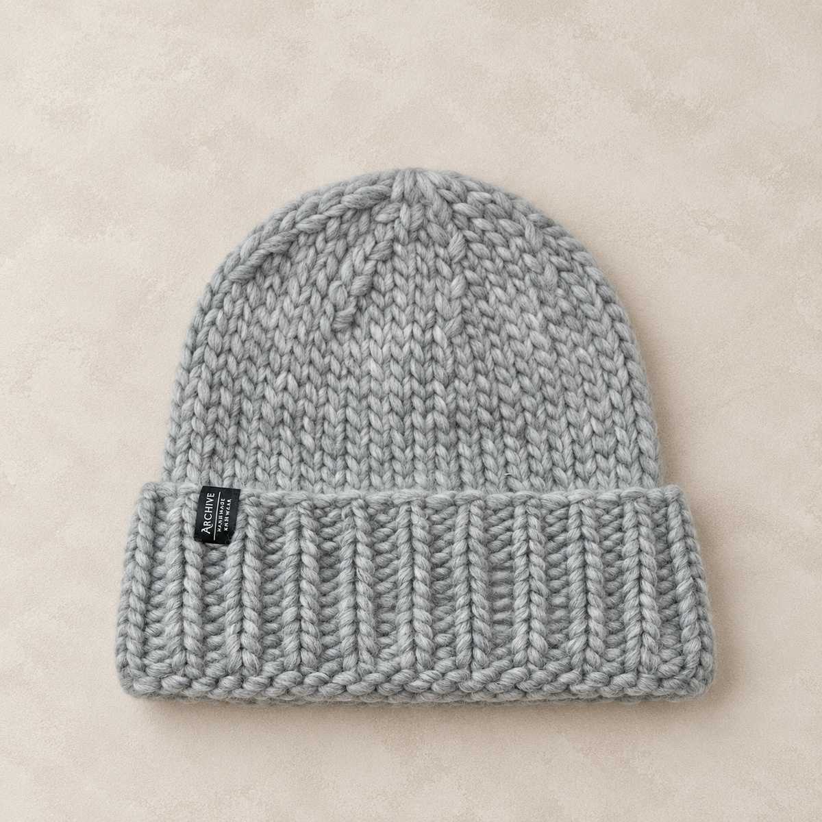 A light gray chunky knit beanie hat with a folded cuff, laid flat on a textured beige background. A small black fabric brand tag is visible on the left side of the cuff.