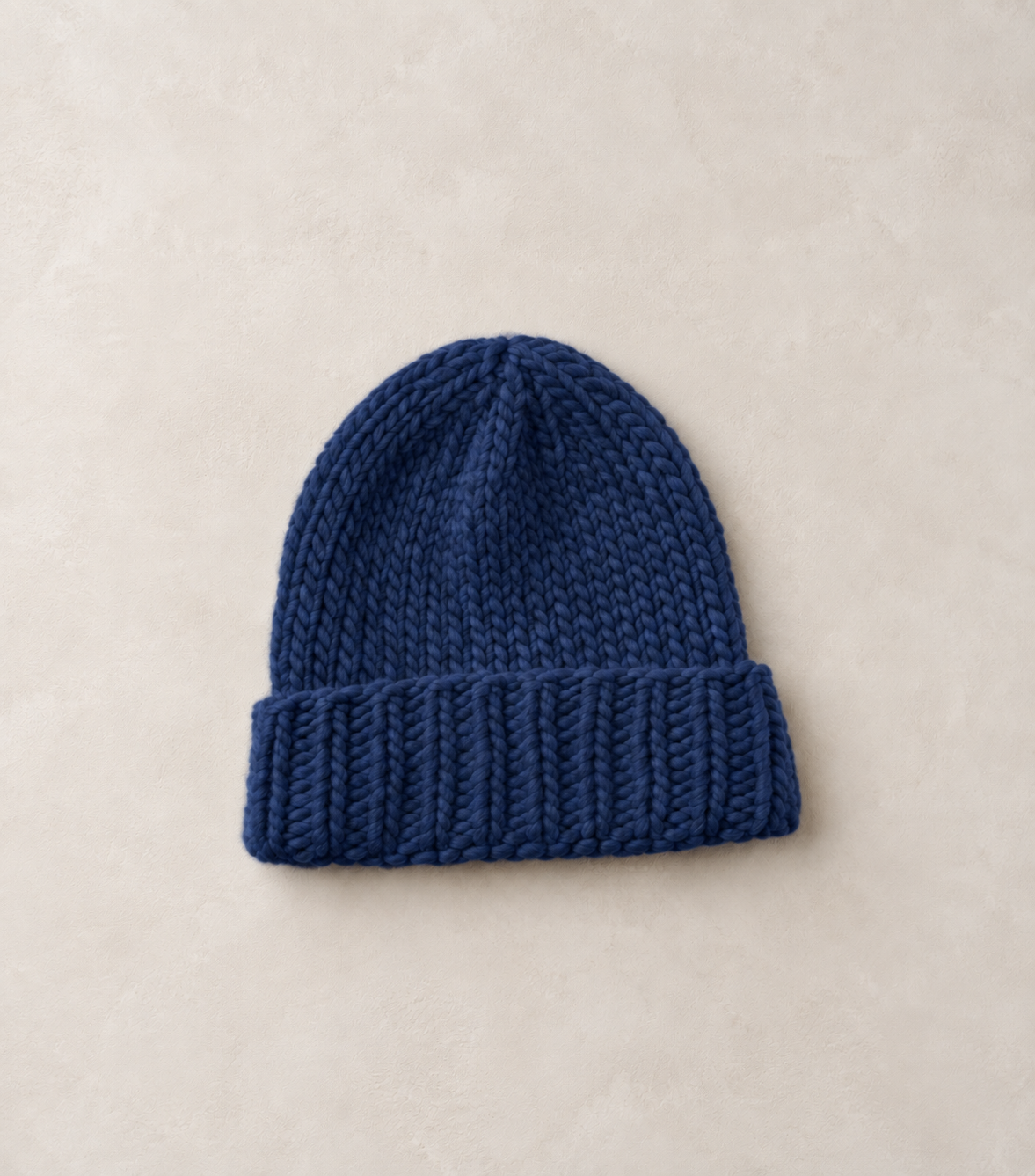 A dark blue chunky knit beanie hat with a folded cuff, displayed flat on a light beige textured background.