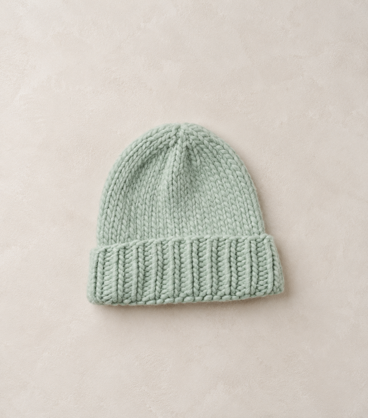A light mint green knit beanie hat with a ribbed cuff, laid flat on a textured beige background.