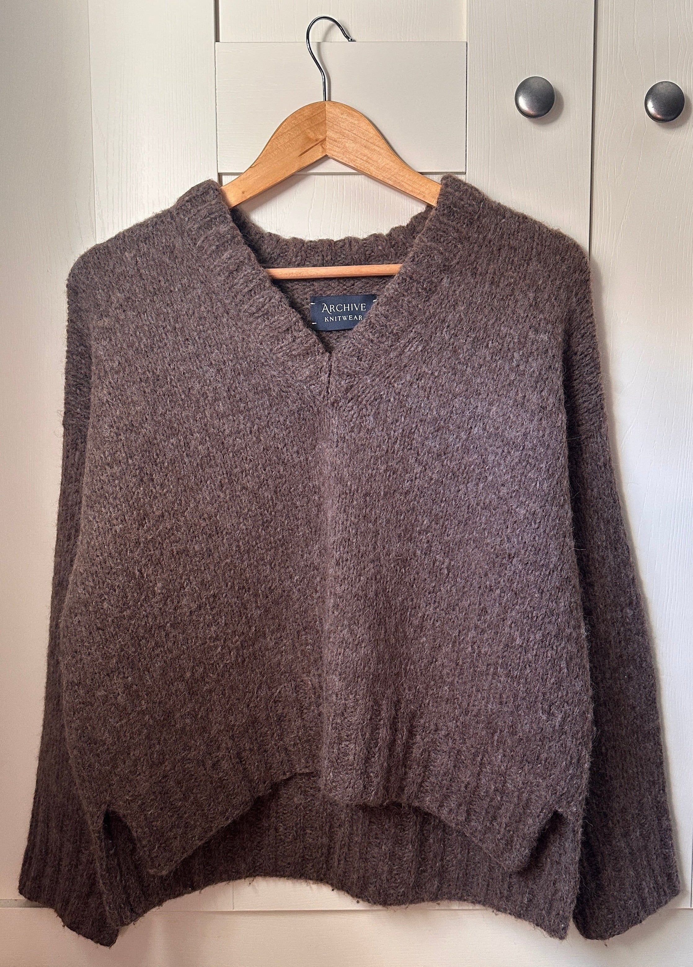 A dark brown knit sweater with a V-neck and long sleeves, hanging on a wooden hanger against a white paneled closet door.