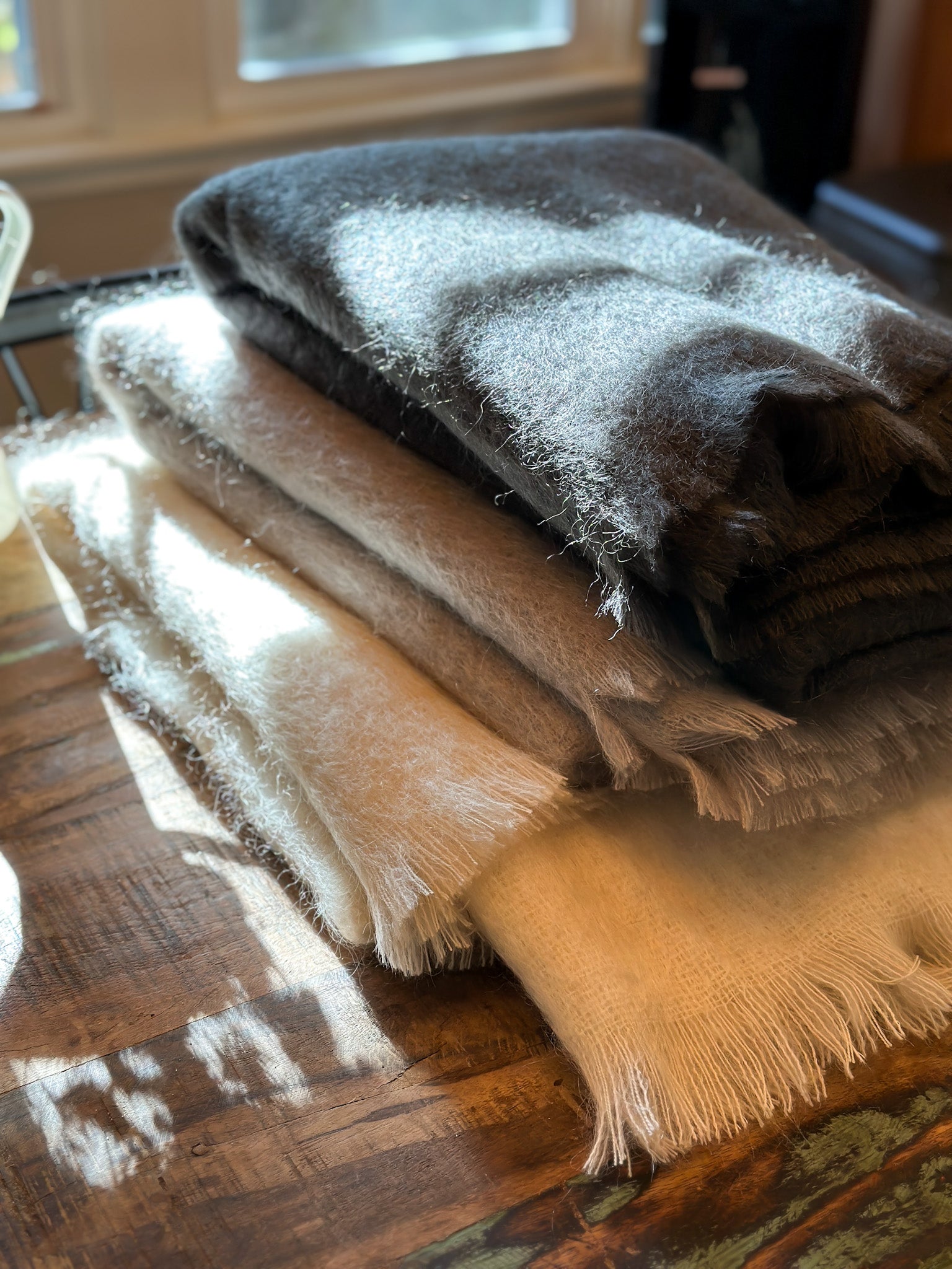 Stack of folded fluffy mohair blankets in brown and beige on a wooden surface with a window in the background.