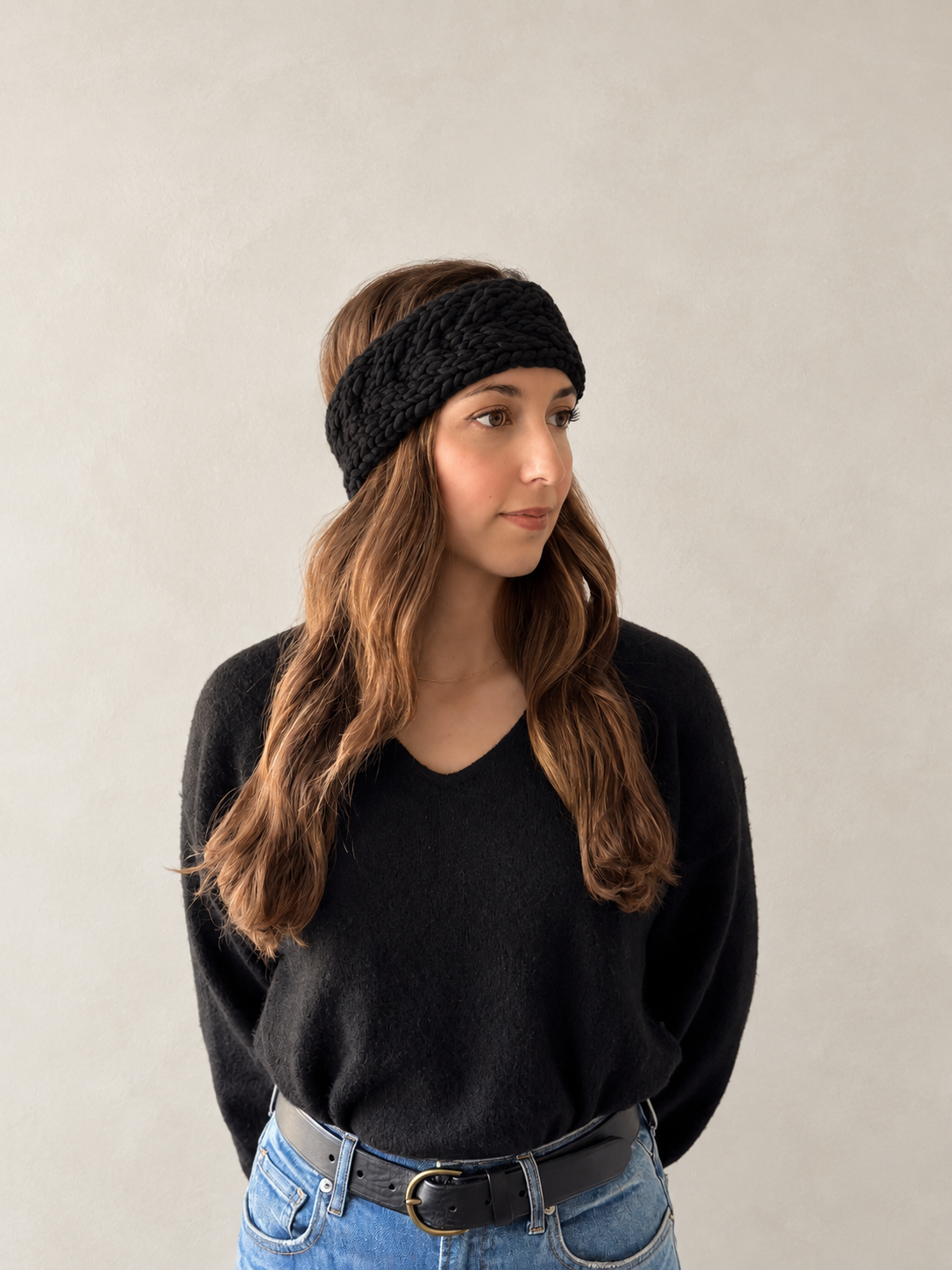 A woman with long brown hair wearing a black textured knit headband, a black V-neck sweater, and blue jeans with a black belt, standing against a neutral light gray wall.