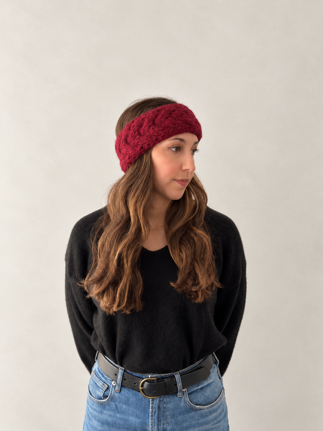 A woman wearing a chunky, cable-knit burgundy headband, styled with a black sweater and blue jeans against a plain light gray wall.
