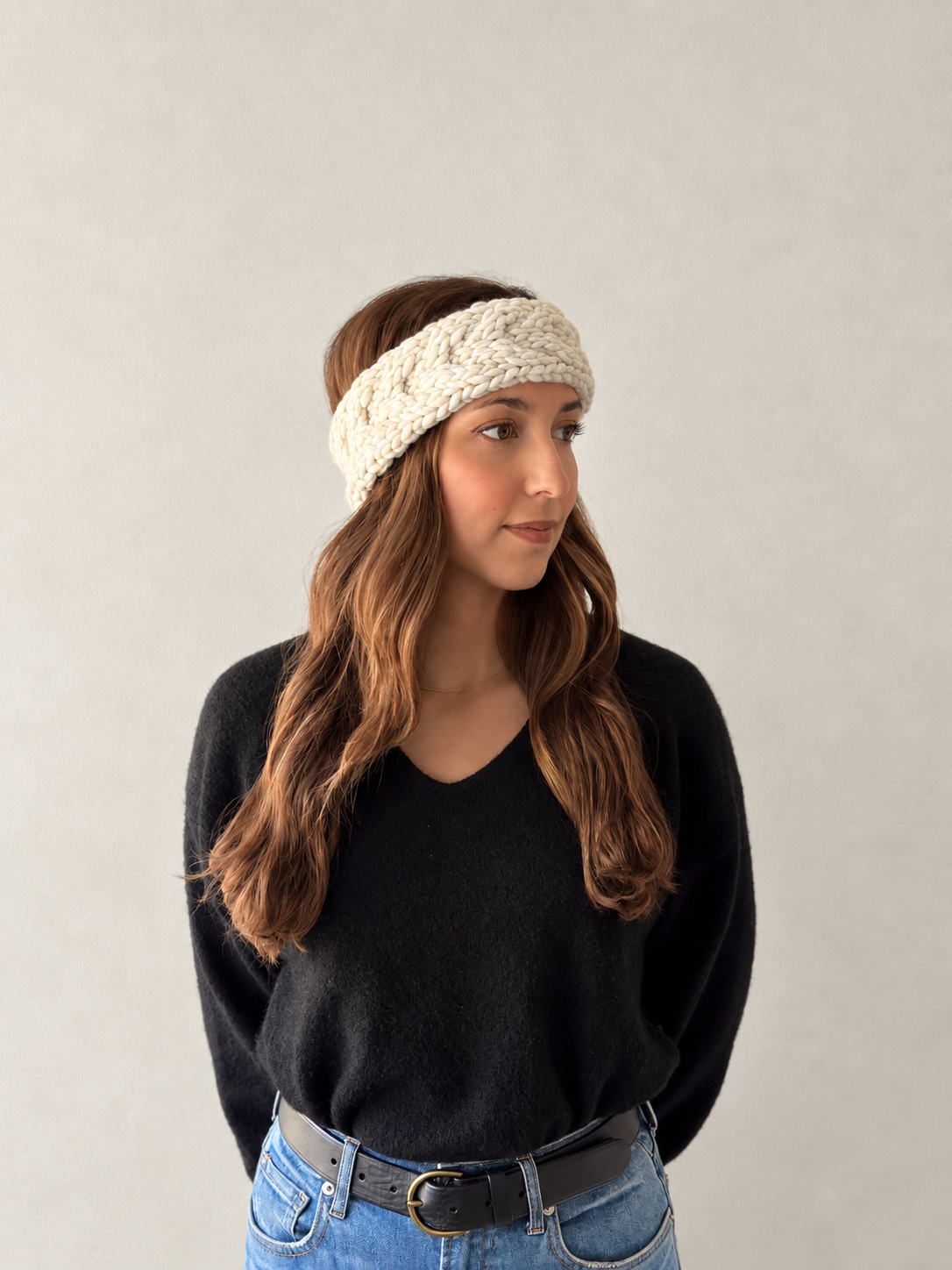 A woman with long brown hair wearing a cream-colored, textured cable-knit headband, a black V-neck sweater, and blue jeans, standing against a neutral light gray background.