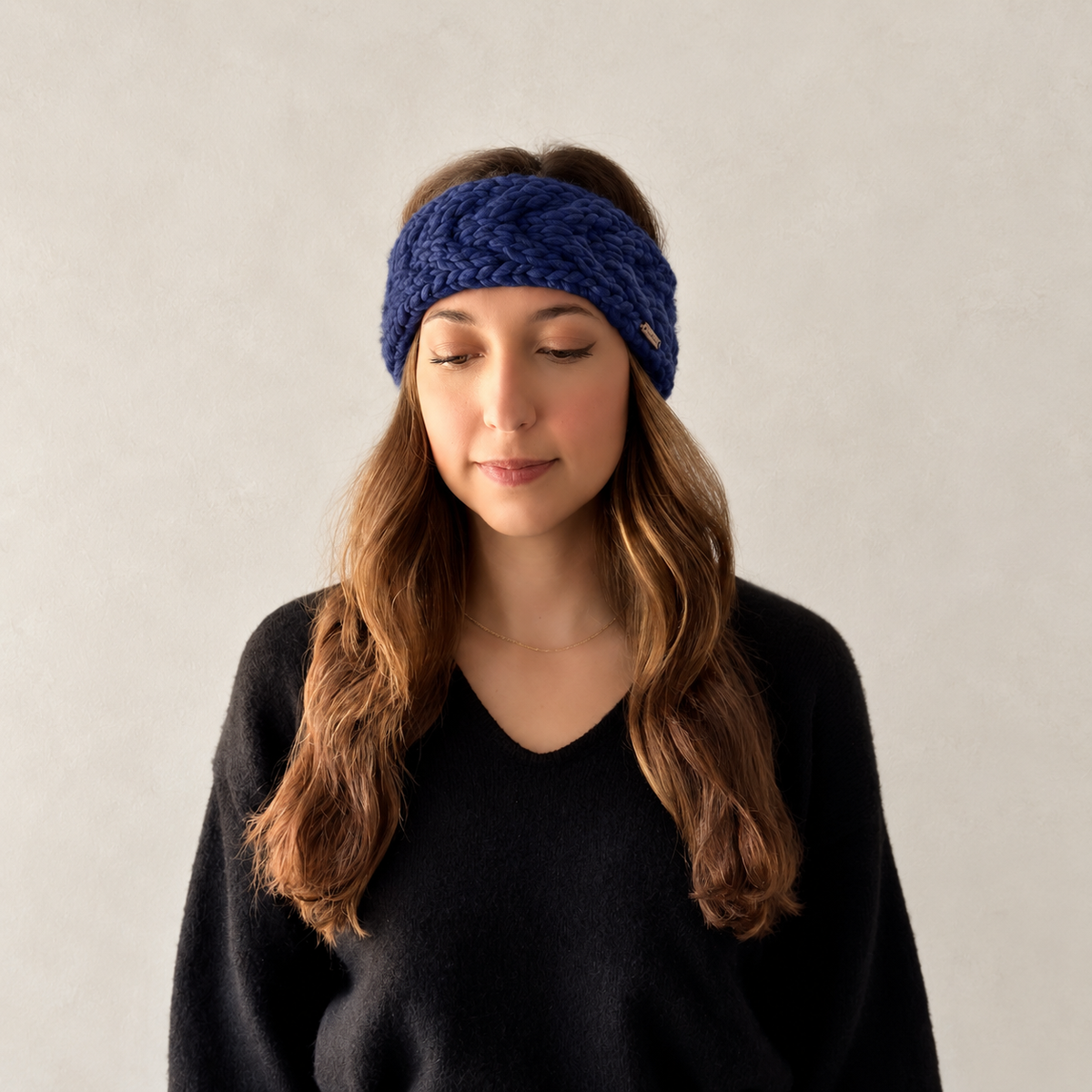 A woman with long brown hair wearing a knitted navy blue cable knit headband and a black sweater, photographed against a neutral background.
