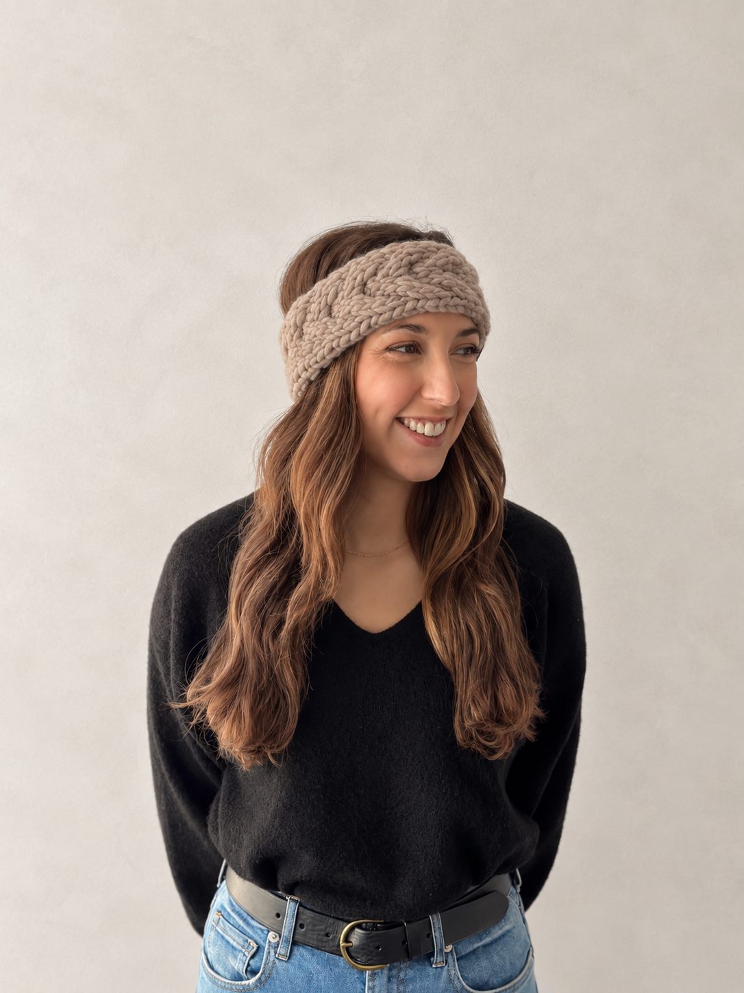 A woman wearing a chunky knit cable-patterned headband in taupe, styled with a black sweater and blue jeans against a neutral textured wall.