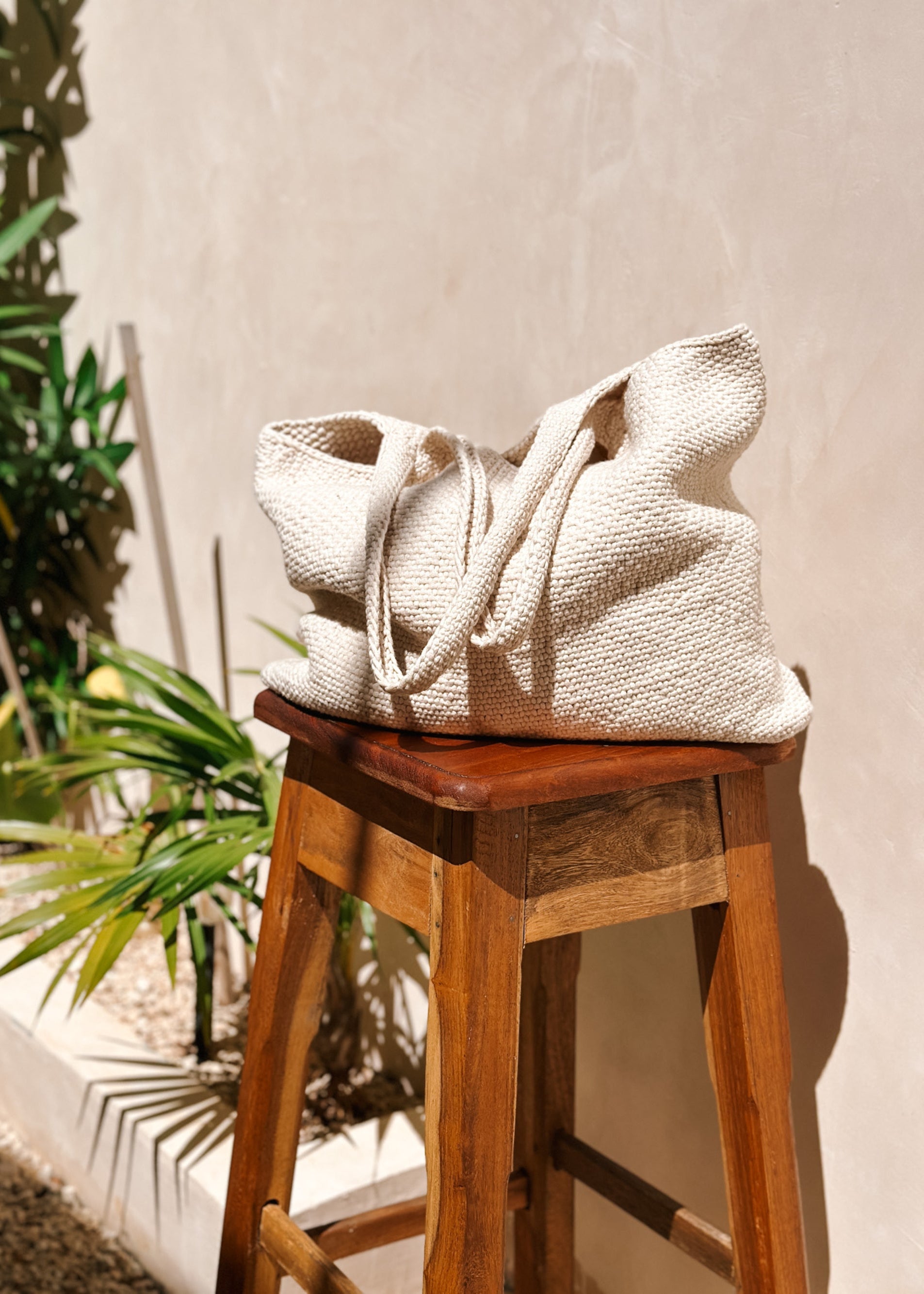 white knitted tote bag sitting on a wooden stool in the sun with plants in background