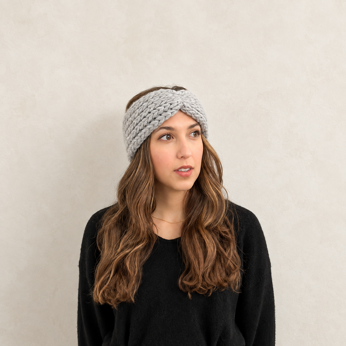 A woman with long wavy brown hair wearing a chunky light gray knitted twisted headband and a black sweater, photographed against a plain light beige wall.