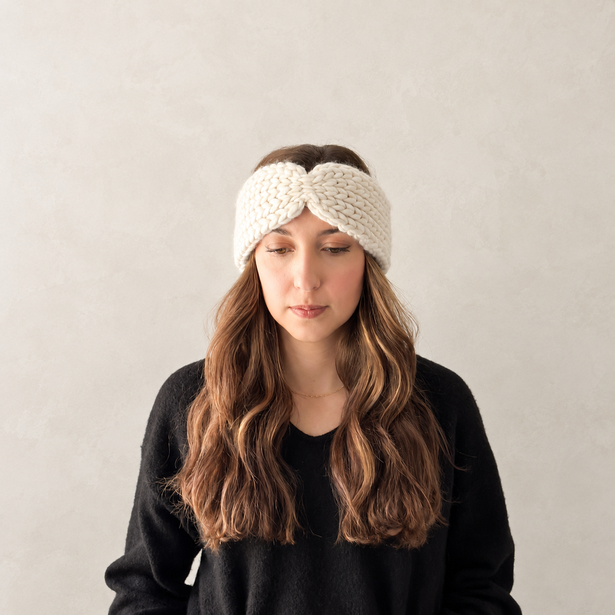 A woman with long wavy brown hair wearing a cream-colored, chunky-knit twisted headband and a black knit sweater, looking down against a neutral light gray textured background.