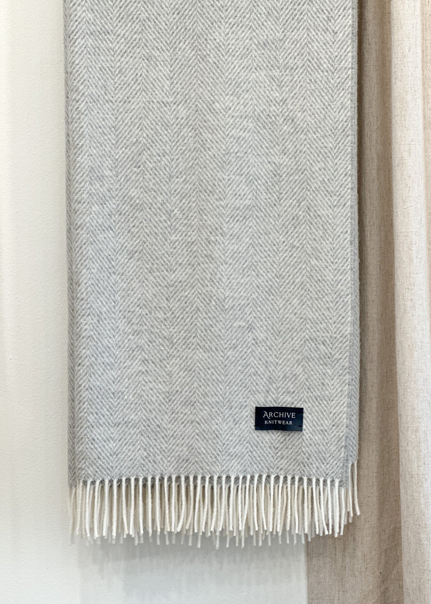 Gray woven blanket with white fringe and herringbone pattern, a brand label on a neutral background