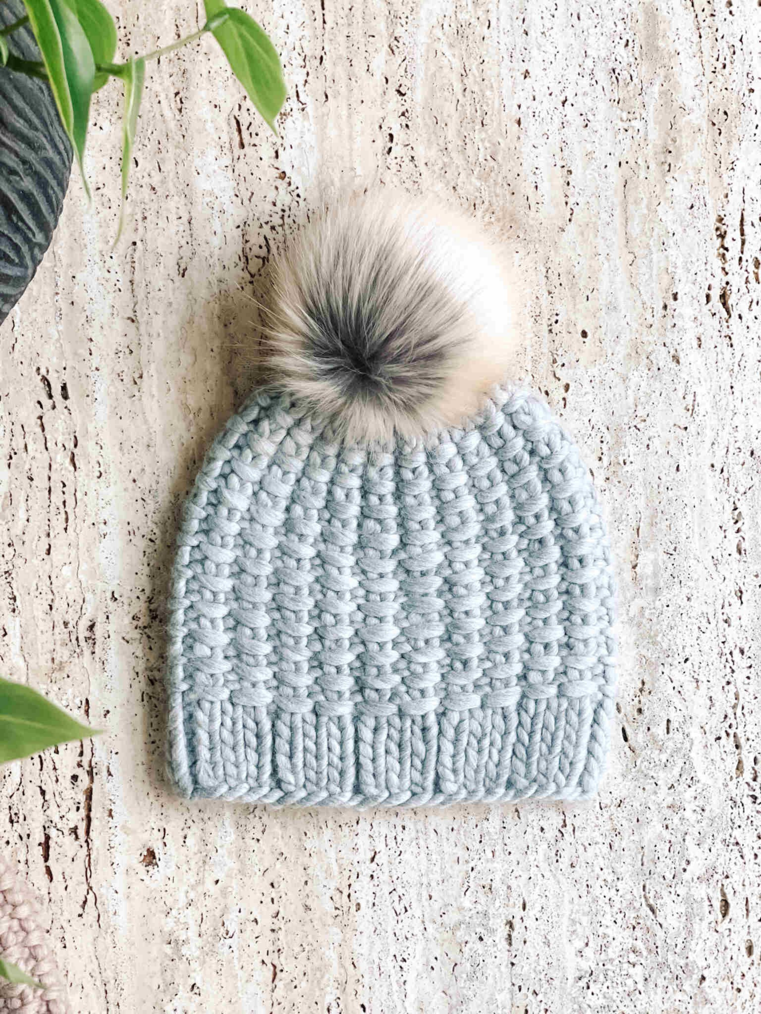 Grey Hand Knit beanie hat topped with faux fur pom pom on a beige textured surface