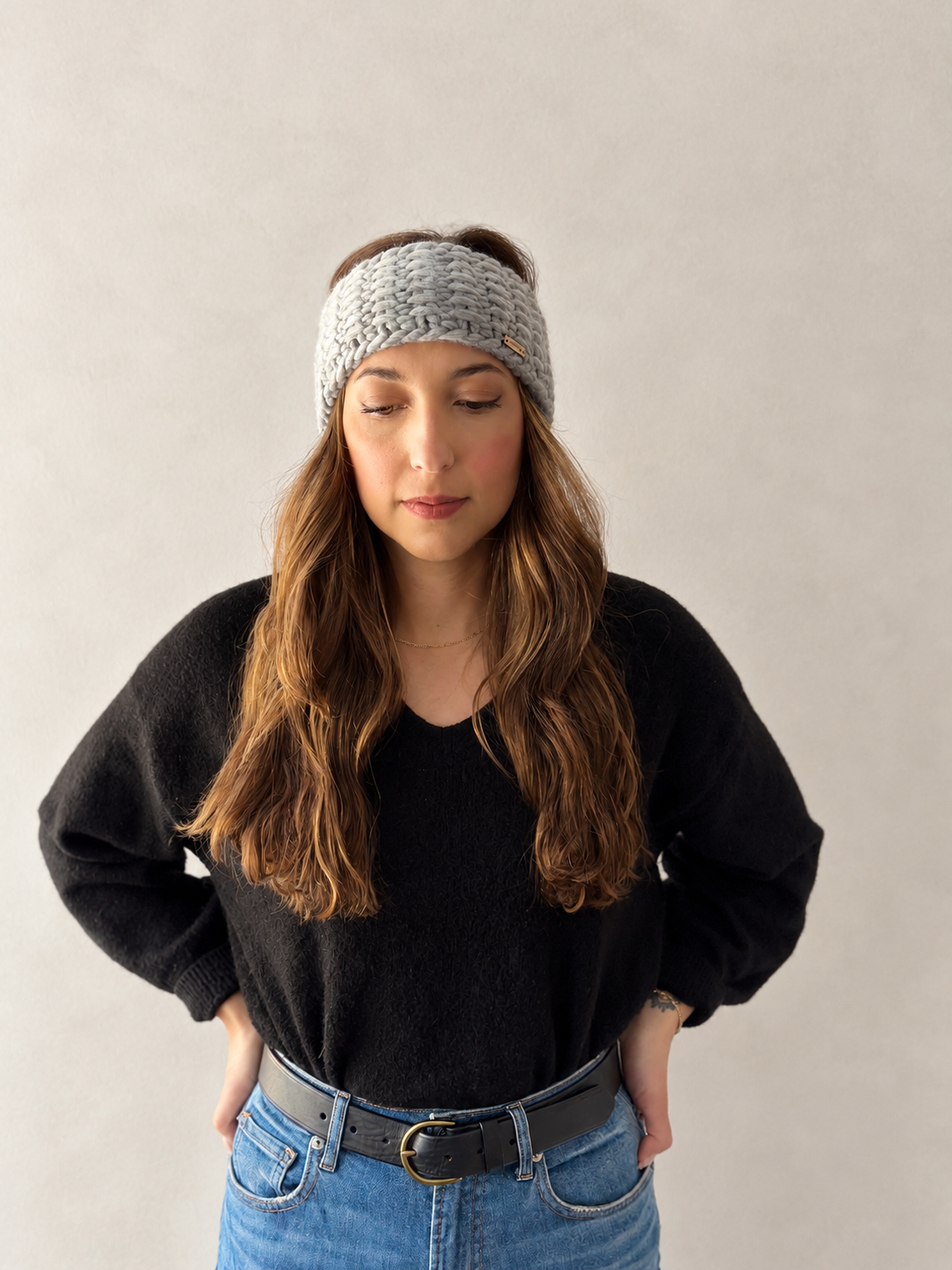 A woman wearing a light gray chunky knit beanie, a black knit sweater, and blue jeans, standing against a neutral light gray background.
