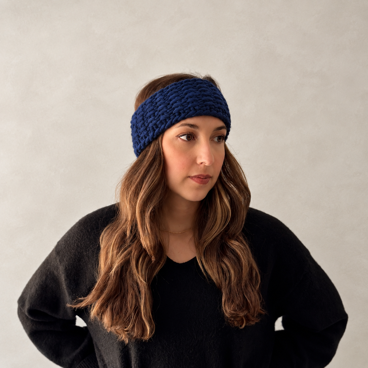 A woman with long wavy brown hair wearing a thick, chunky navy blue knit headband and a black sweater, photographed against a neutral light gray background.