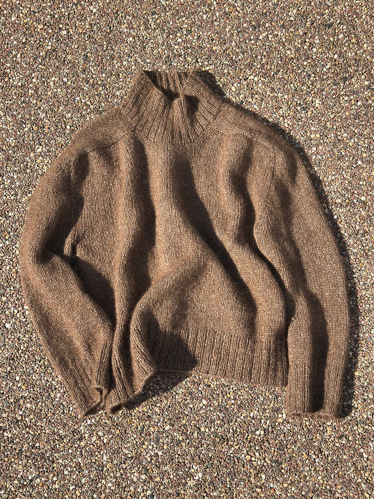 A brown knit turtleneck sweater laid flat on a textured gravel surface.