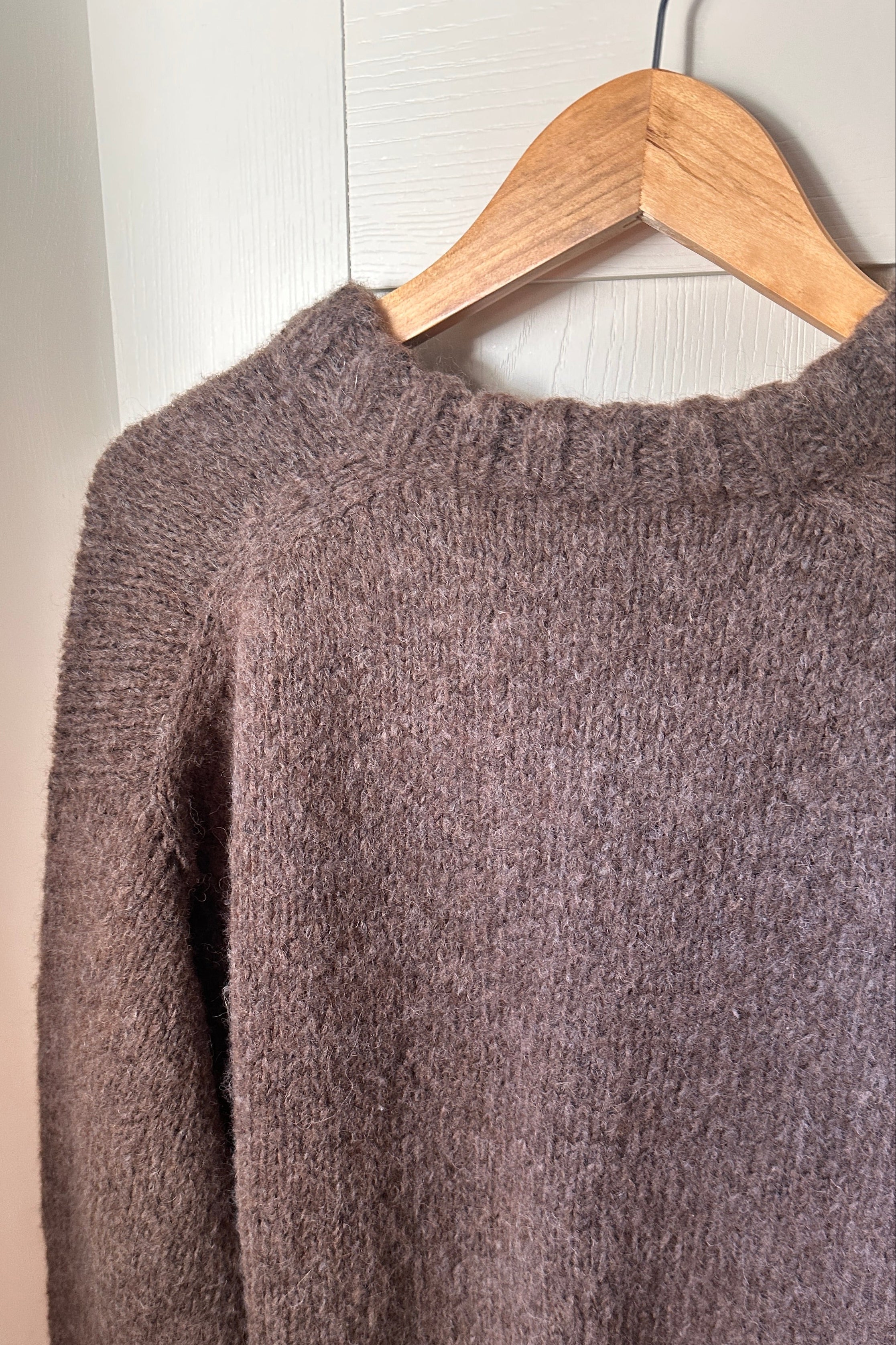 A fuzzy knit crewneck sweater in a muted taupe-brown color, hanging on a wooden hanger against a white paneled door.