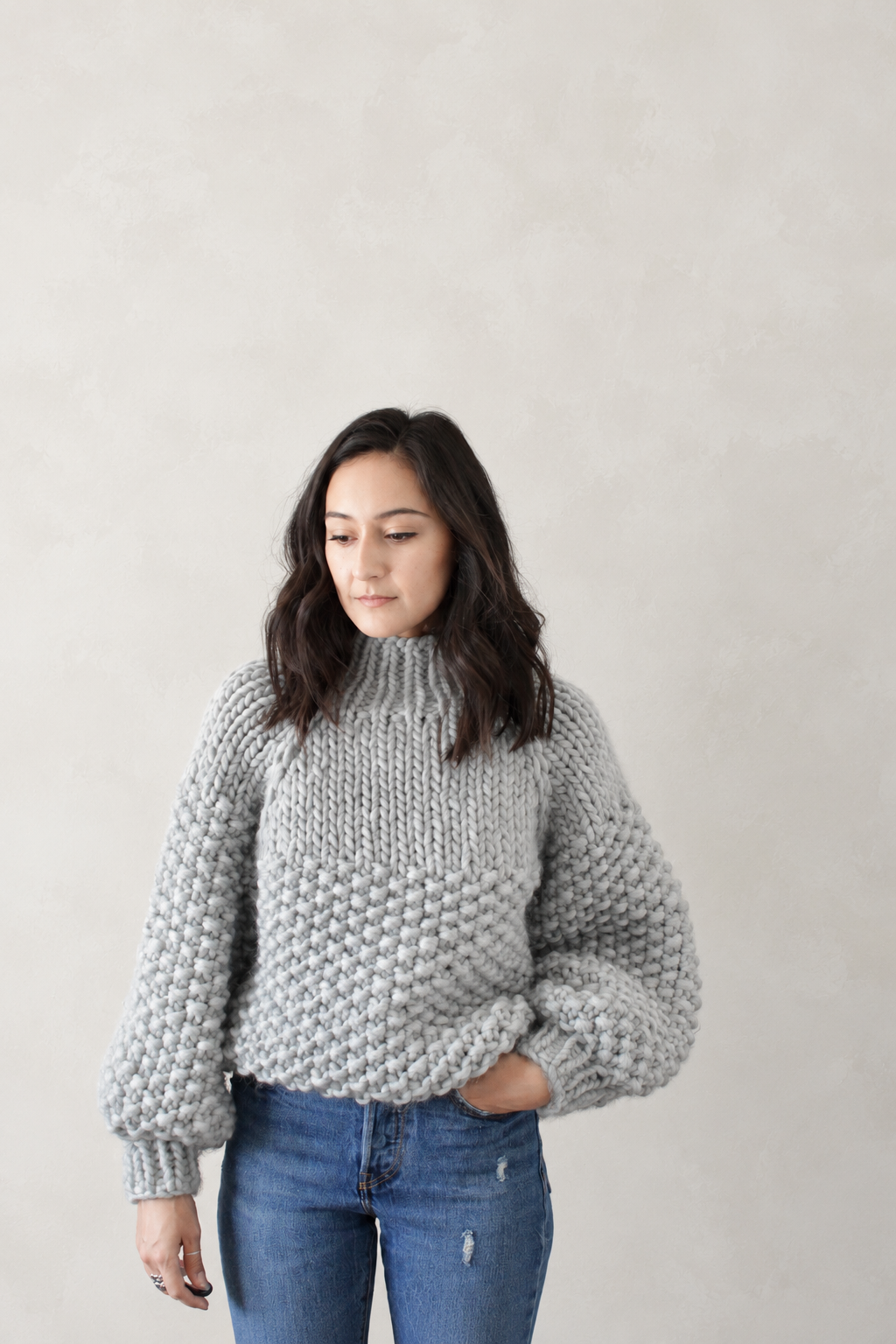 A woman wearing a chunky light gray knit mock-neck sweater with balloon sleeves and blue jeans, standing against a neutral textured beige wall.