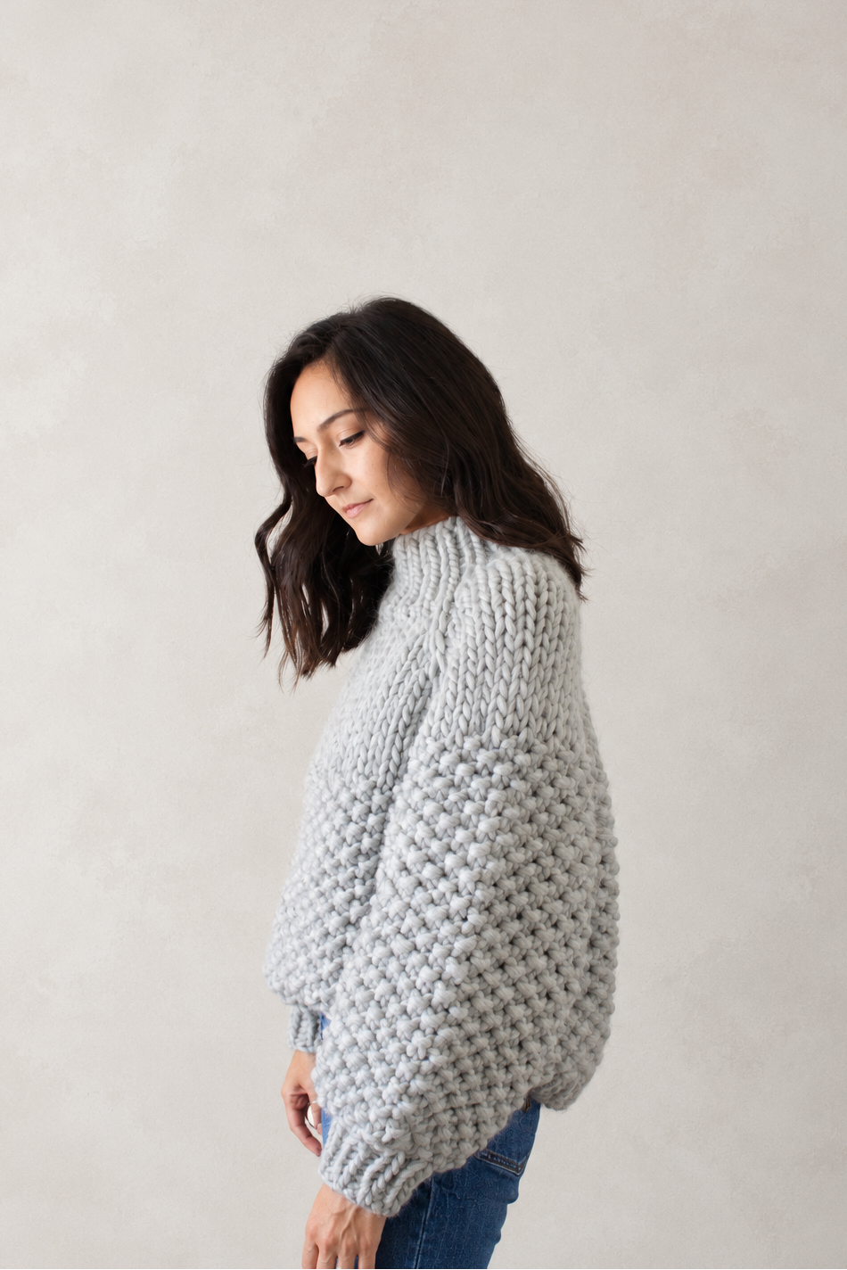 A woman wearing a chunky light gray knit sweater and blue jeans, standing against a neutral textured beige wall.